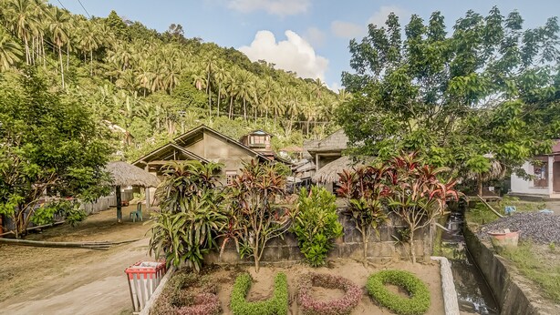 Un paysage rural avec une maison entourée d’une végétation luxuriante et une colline en arrière-plan.