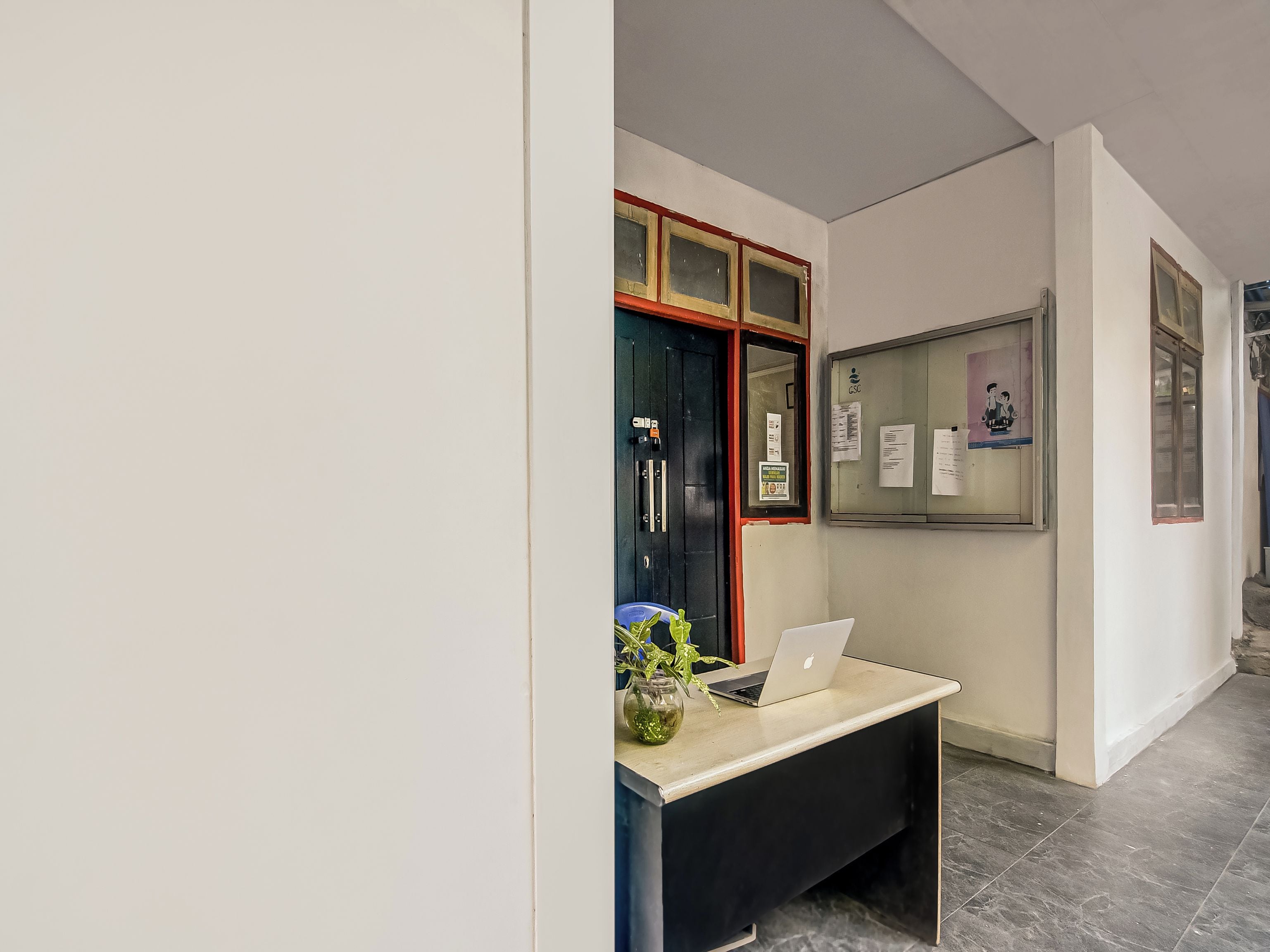 A reception desk with a laptop, a vase with green plants, and a bulletin board with papers.