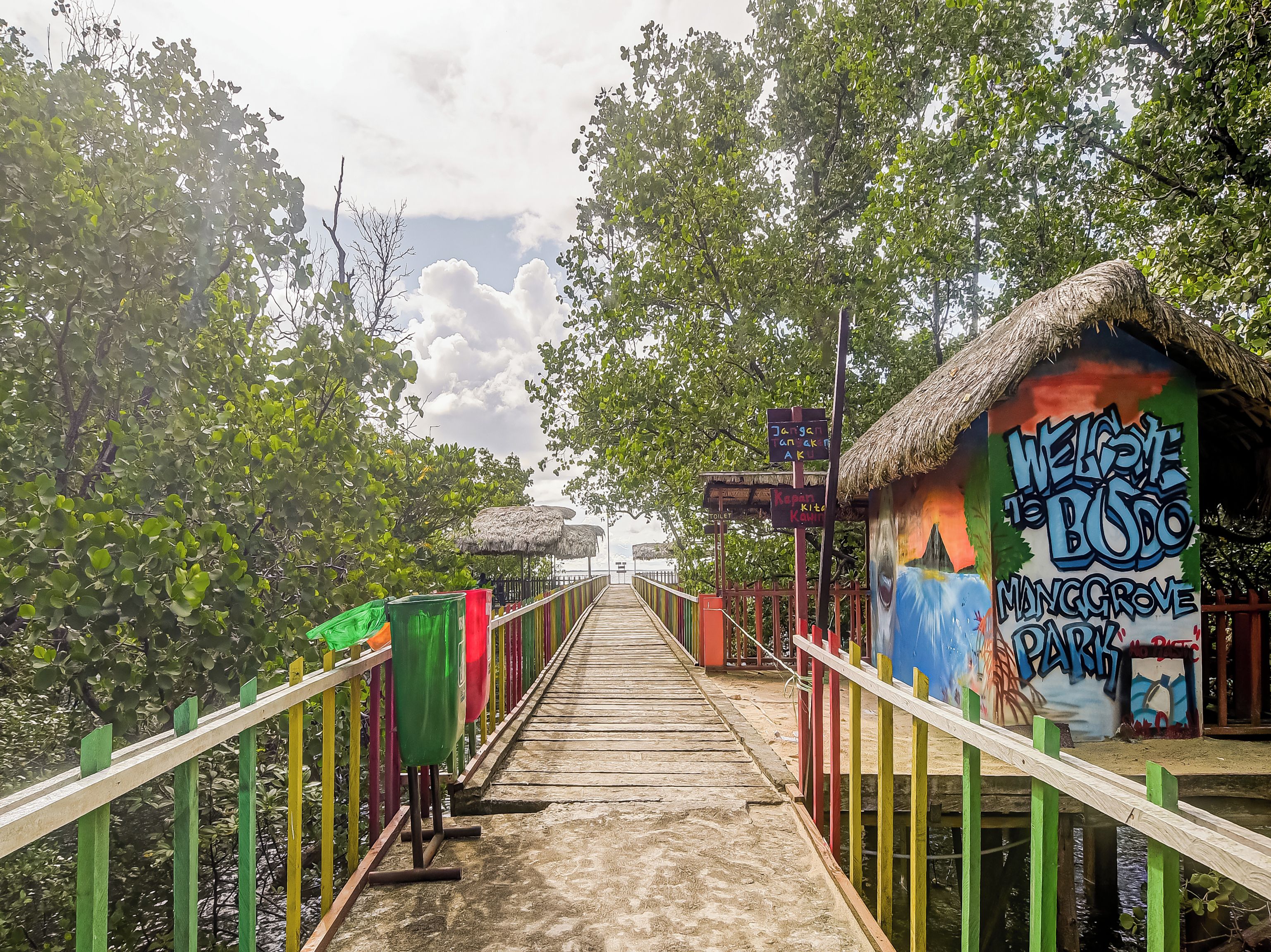 Un pasillo de madera que conduce a una estructura de techo de paja con un mural colorido y un cartel.
