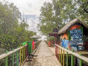 A wooden walkway leading to a thatched-roof structure with a colorful mural and a signboard. - OYO Homes 91118 Homesstay Desa Wisata Budo (Wori)