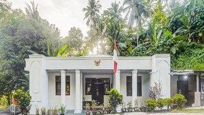 Un bâtiment à la façade blanche, un mât sur lequel flotte un drapeau, et une rangée de plantes en pot devant l’entrée.