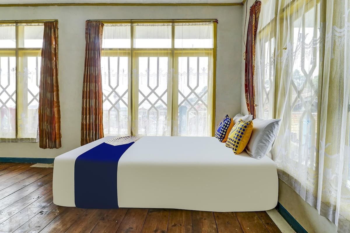 A bed in a room with large windows, wooden flooring, and a view of buildings outside.