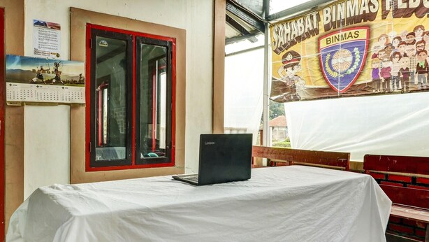 Una habitación con una mesa cubierta por un mantel blanco, una laptop sobre la mesa, un ventanal con marco rojo, y una pared con un calendario y un cartel.