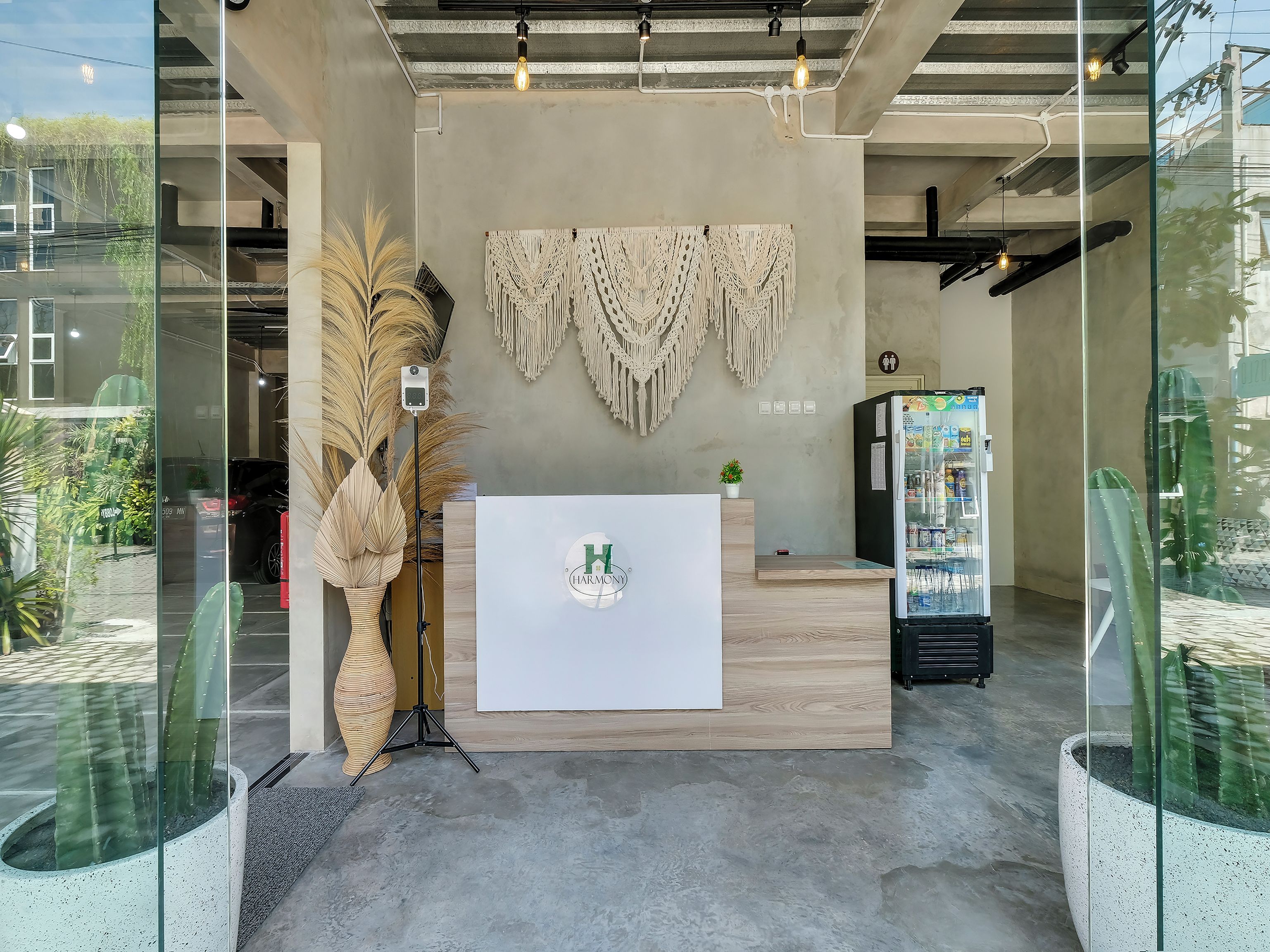 A reception area with a wooden reception desk, a wall-mounted macrame decoration, a refrigerator, and potted plants.