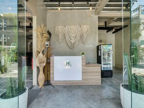 Et receptionsområde med en træreception, en vægophængt makramevævning, et køleskab og potteplanter.