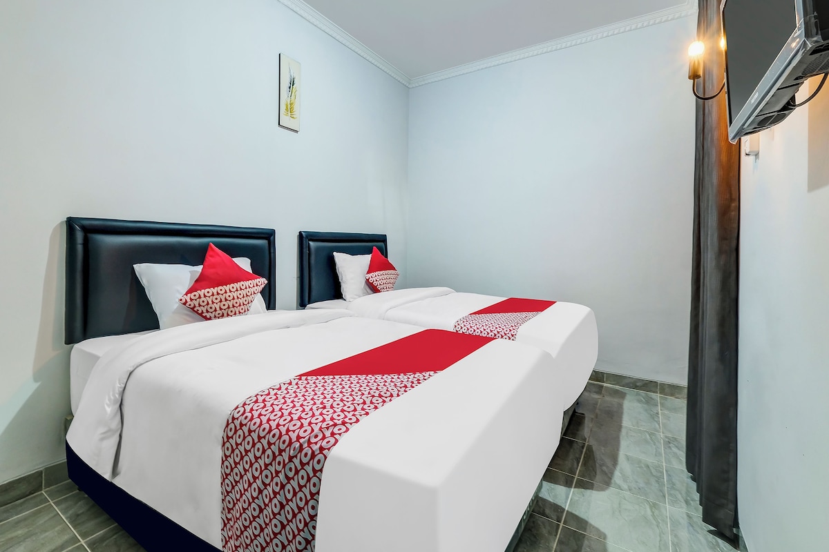 Ein Hotelzimmer mit einem Bett, zwei Kissen, einer rot-weißen Bettdecke, einem an der Wand montierten Fernseher und einer Deckenleuchte.