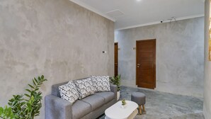 A modern living room with a grey sofa, a white coffee table, and two grey ottomans. There are potted plants and a wooden door in the background. - OYO Flagship 90983 Harmony Easy Stay (Depok)