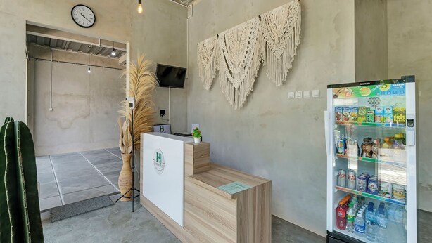 A reception area with a wooden desk, a clock, a macrame wall hanging, and a beverage refrigerator stocked with drinks. - OYO Flagship 90983 Harmony Easy Stay (Depok)