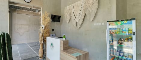 Zona de recepción con escritorio de madera, un reloj, un colgador de macramé y un refrigerador de bebidas lleno.