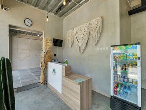 A reception area with a wooden desk, a clock, a macrame wall hanging, and a beverage refrigerator stocked with drinks. - OYO Flagship 90983 Harmony Easy Stay (Depok)