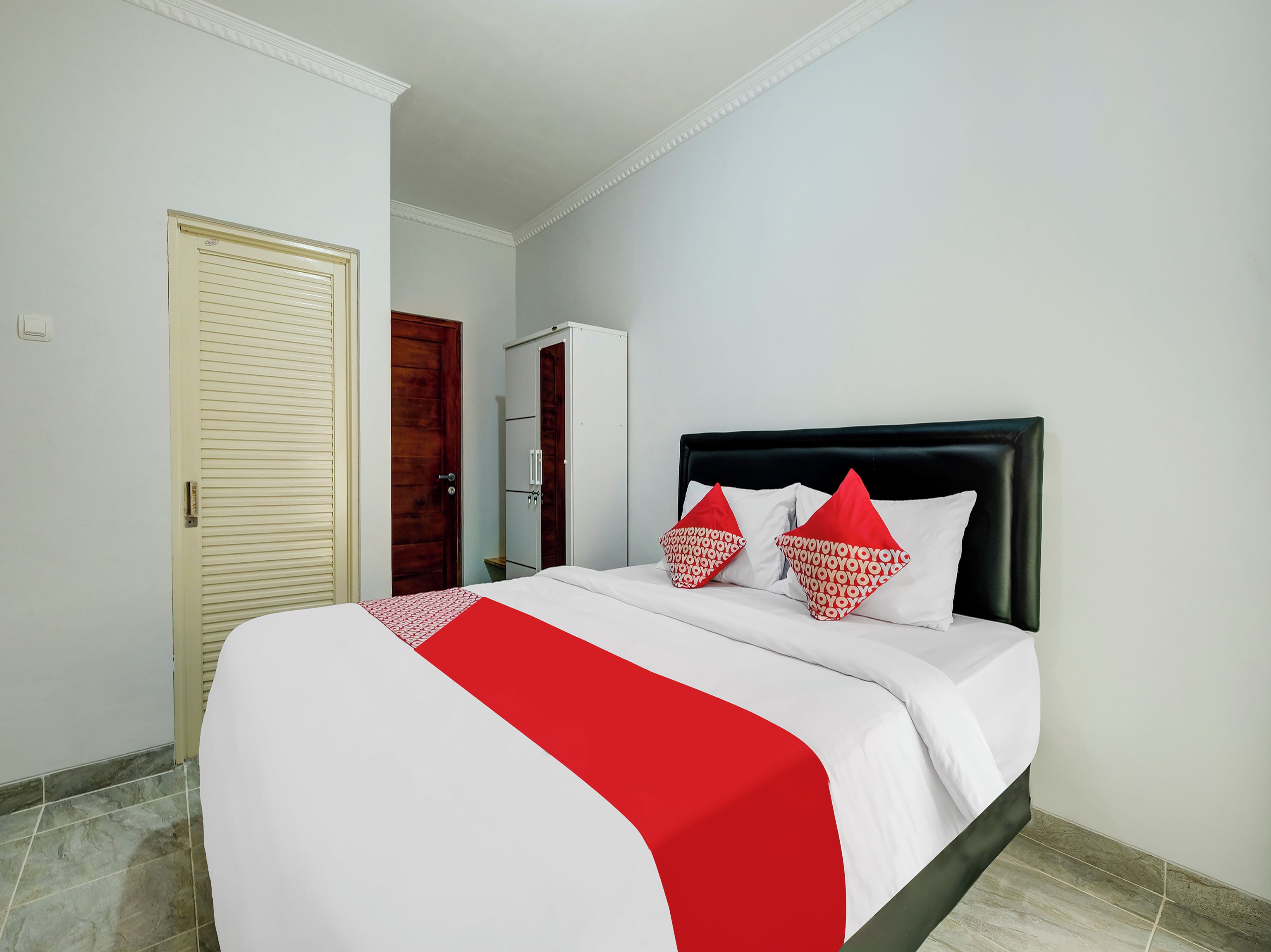 A neatly made bed with a red and white bedspread, two red pillows, and a dark headboard in a room with white walls and a tiled floor.