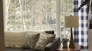 A living room with a dark sofa, patterned cushions, a small table with a lamp, and a view through large windows with a blue and white checkered curtain. - Waterfront Cottage| Firepit Family Retreat 10mins to DT Huntsville & Deerhurst (Huntsville)