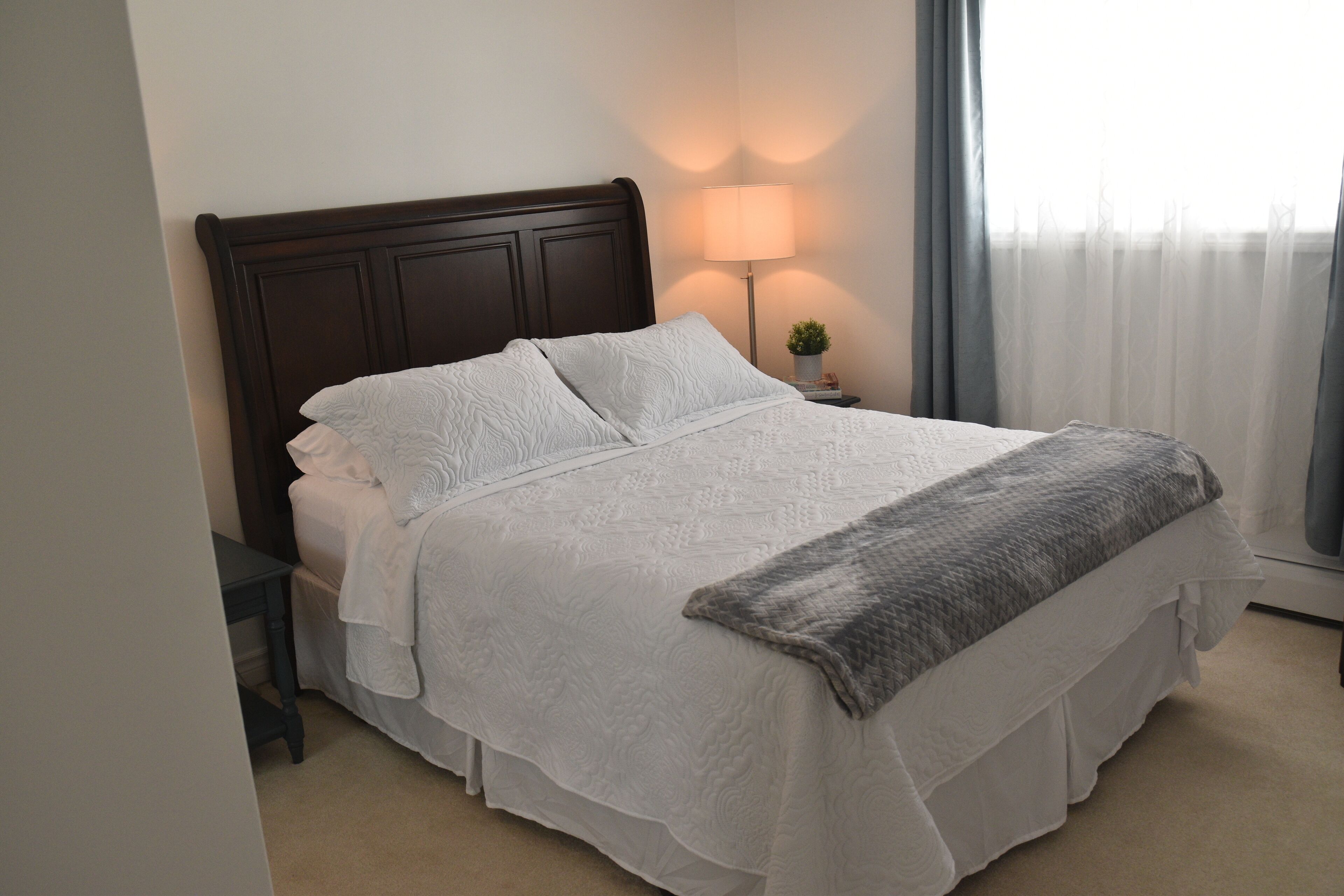 A neatly made bed with white linens and a gray throw blanket, a bedside lamp, and a small potted plant on a nightstand.