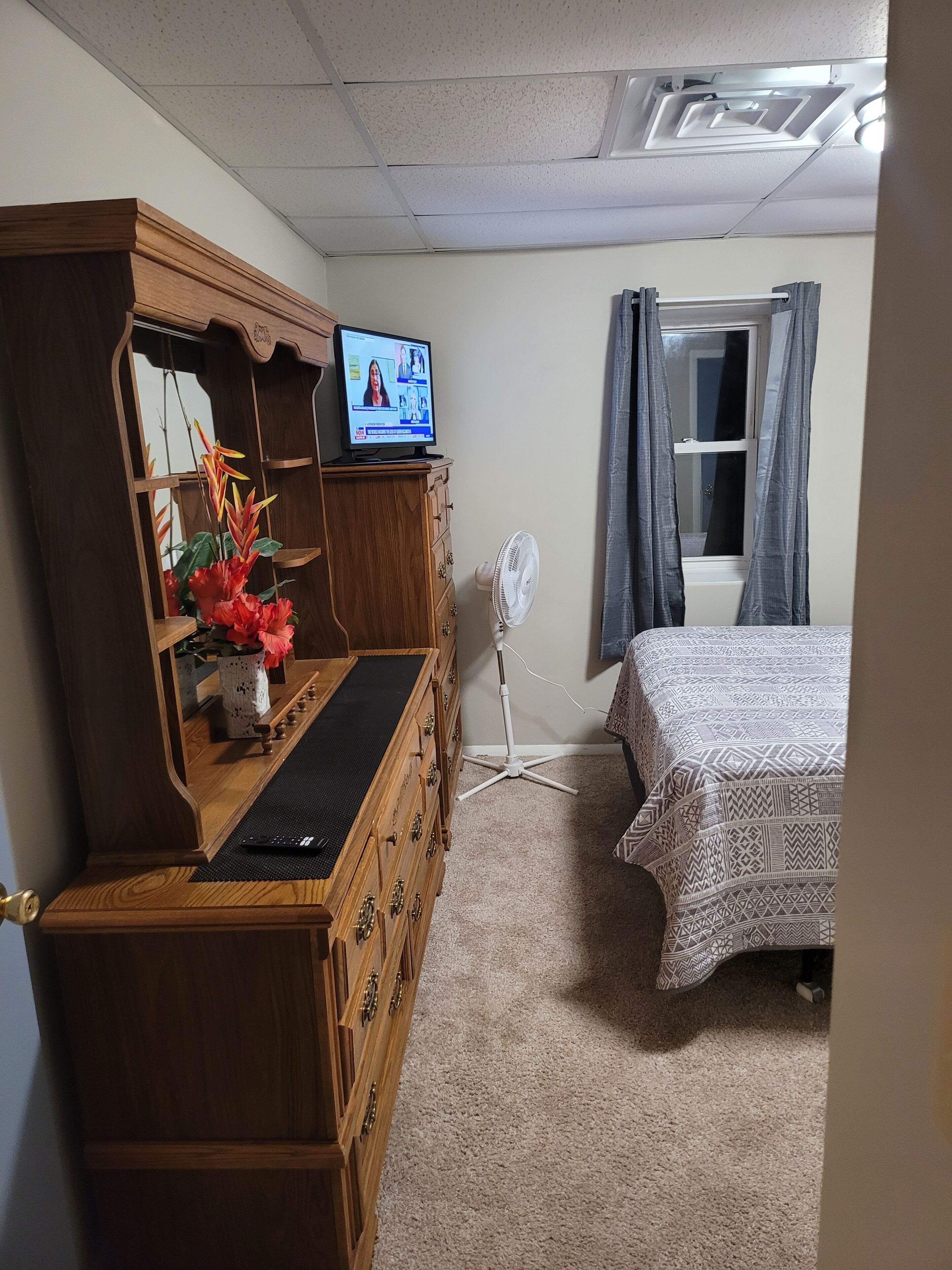Une chambre avec une commode en bois, un téléviseur posé dessus, un lit avec une literie à motifs, un ventilateur sur pied et une fenêtre avec des rideaux.