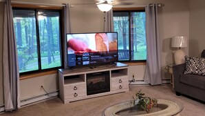 Un soggiorno con televisione su un supporto in legno, una ventola a soffitto e vista su una foresta attraverso ampie finestre.