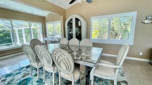 A dining area with a marble table and white chairs, large windows with blinds, and a view of greenery outside.