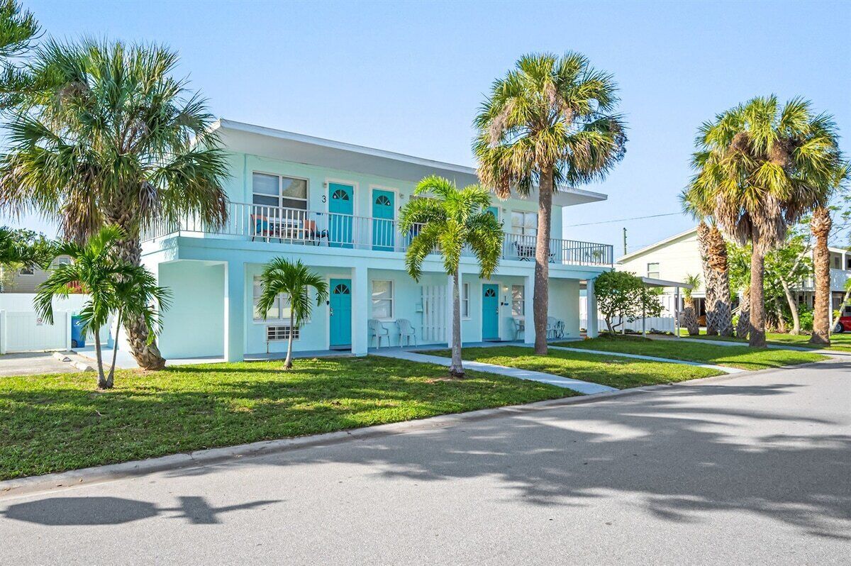A two-story motel with a light blue exterior, multiple balconies, and palm trees in the front yard.