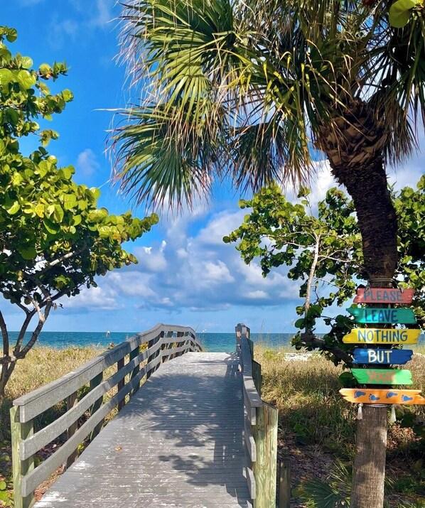 A wooden boardwalk leading to a beach with a palm tree and a signpost with various messages.