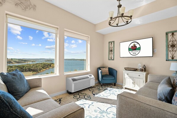 A living room with a large window offering a scenic view of a lake and mountains, a chandelier, and a framed logo on the wall. - Lakeview Penthouse 4BR/4BA Condo • Sleeps 12 • Sunroom + Balcony (Branson)