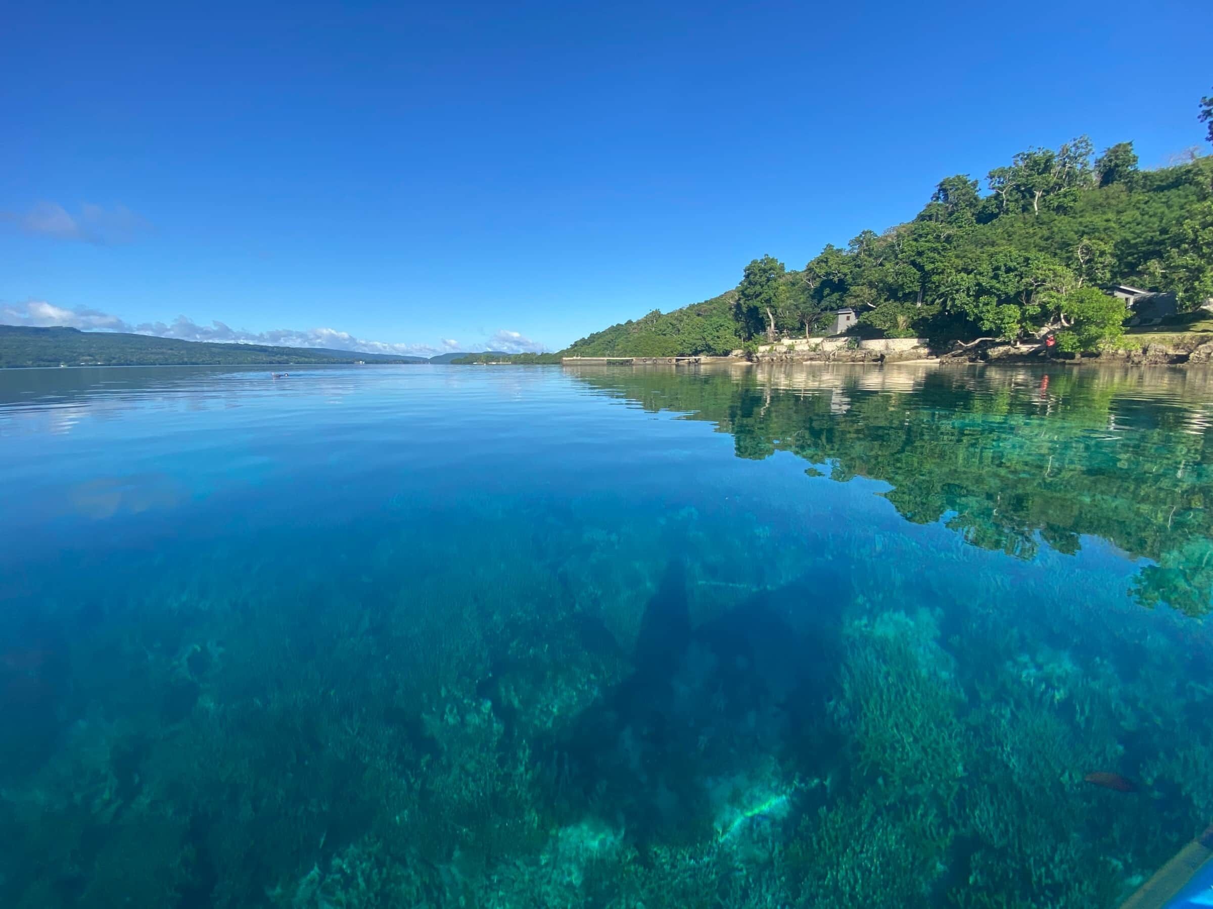 A calm lake with clear water reflecting the surrounding landscape, including a forested hillside and a small building.