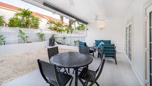 Eine überdachte Terrasse mit rundem Tisch und Stühlen, einem Deckenventilator und Blick auf ein Gebäude mit rotem Dach sowie viel Grün im Hintergrund.
