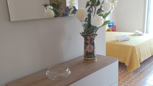 Une chambre d’hôtel avec une console en bois, un vase de fleurs, un lit recouvert d’une courtepointe jaune et un sol carrelé.