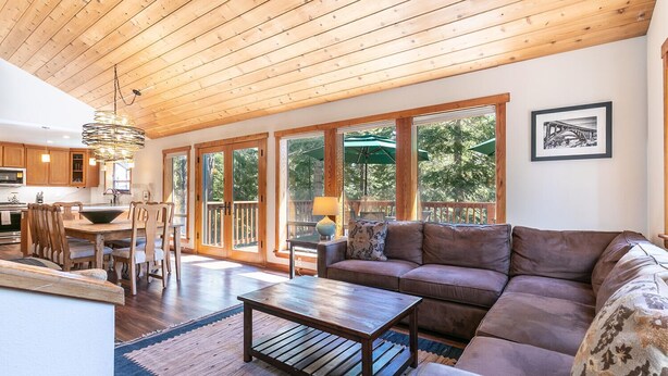 A spacious living room with a wooden ceiling, large windows, a sofa, and a dining area with wooden furniture.