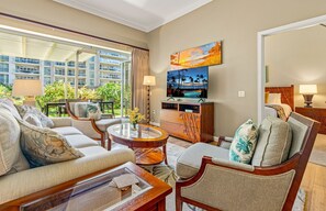 A hotel room with a sofa, armchair, coffee table, TV, and a view of the hotel building and greenery.