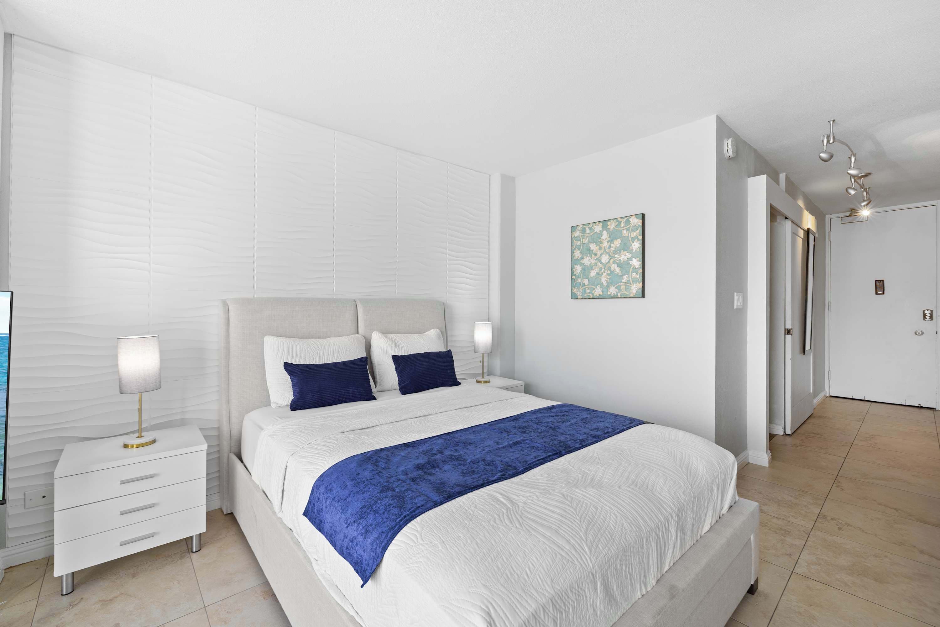 Ein Schlafzimmer mit einem großen Bett, weißen Nachttischen, einer blauen Tagesdecke und einer Wandlampe.
