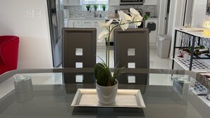 Una cocina moderna con armarios blancos, un refrigerador de acero inoxidable y un microondas. Hay un comedor con una mesa de vidrio y sillas, una planta en maceta y un pequeño estante con botellas.
