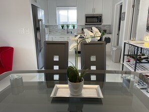 A modern kitchen with white cabinets, a stainless steel refrigerator, and a microwave. There is a dining area with a glass table and chairs, a potted plant, and a small shelf with bottles. - Clean and modern 2BR/2Bath Jacuzzi home, beautiful Patio, minutes from the beach (Fort Lauderdale)