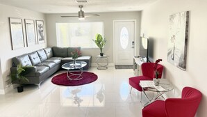 Una sala de estar moderna con un sofá seccional gris, una mesa de centro de vidrio, un sillón rojo y una alfombra roja. Se observa una puerta con panel de vidrio, un ventilador de techo y obras de arte enmarcadas en las paredes.