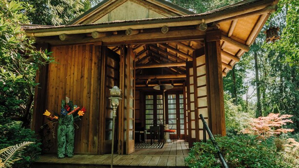 A wooden cabin with a porch, surrounded by lush greenery and a stone staircase leading up to it.