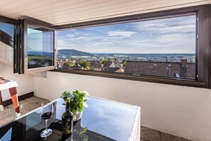Een kamer met een groot raam met uitzicht op een stad en heuvels. Er staat een tafel met een fles en glas, en een plant erop.