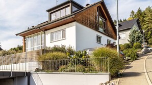 Una casa moderna de dos pisos con techo inclinado, ventanas grandes y un garaje visible.