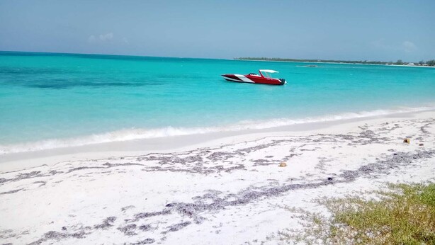 A beach with clear turquoise water, a white sandy shore, and a red and white boat in the distance. - 960 Sq. ft  Beach  Front Villa located in Steventon, Exuma,  Bahamas . (Steventon)