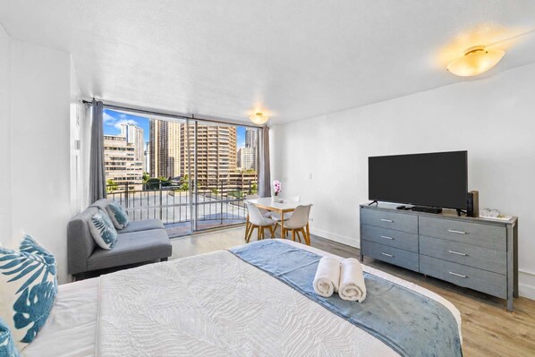 A modern hotel room with a large bed, a desk, a television, and a balcony with a city view.