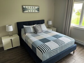 Une chambre à coucher comprenant un lit, deux tables de chevet, une commode, une fenêtre avec des rideaux et un tableau décoratif au mur.