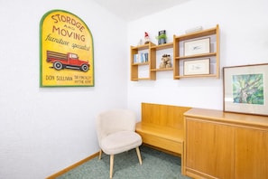 Una habitación con un banco de madera, una silla, un estante en la pared con libros y un osito de peluche, y un cuadro enmarcado.