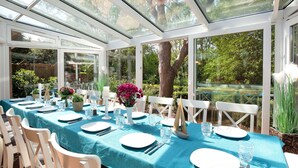 A conservatory with a table set for dining, surrounded by white chairs, and large glass windows offering views of greenery. - Beautiful private villa for 20 people with WIFI, TV, terrace and pets allowed (Bremerhaven)