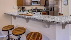 Une cuisine dotée d’un plan de travail en granit, de tabourets en bois, d’appareils électroménagers en acier inoxydable et de luminaires suspendus.