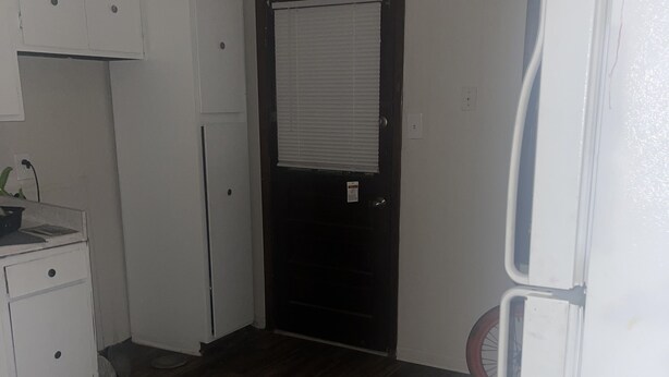 A small, sparsely furnished room with a dark wooden floor, white cabinets, a black door, and a white refrigerator. - BK to JAX CITY GIRL —-> COUNTRY GIRL (Jacksonville)