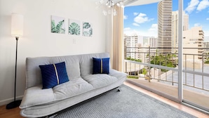 A modern living room with a grey sofa, two blue cushions, a floor lamp, and a chandelier. There are three framed leaf artworks on the wall. - Waikiki Escape – Perfect Location, Near the Beach! (Honolulu)