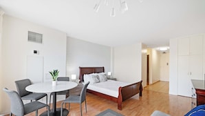 A modern bedroom with a wooden bed, a round dining table with chairs, and a small kitchen area. - Waikiki Escape – Perfect Location, Near the Beach! (Honolulu)