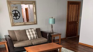 A living room with a sofa, a coffee table, and a chair. There is a lamp on a side table, a mirror on the wall, and a door in the background.