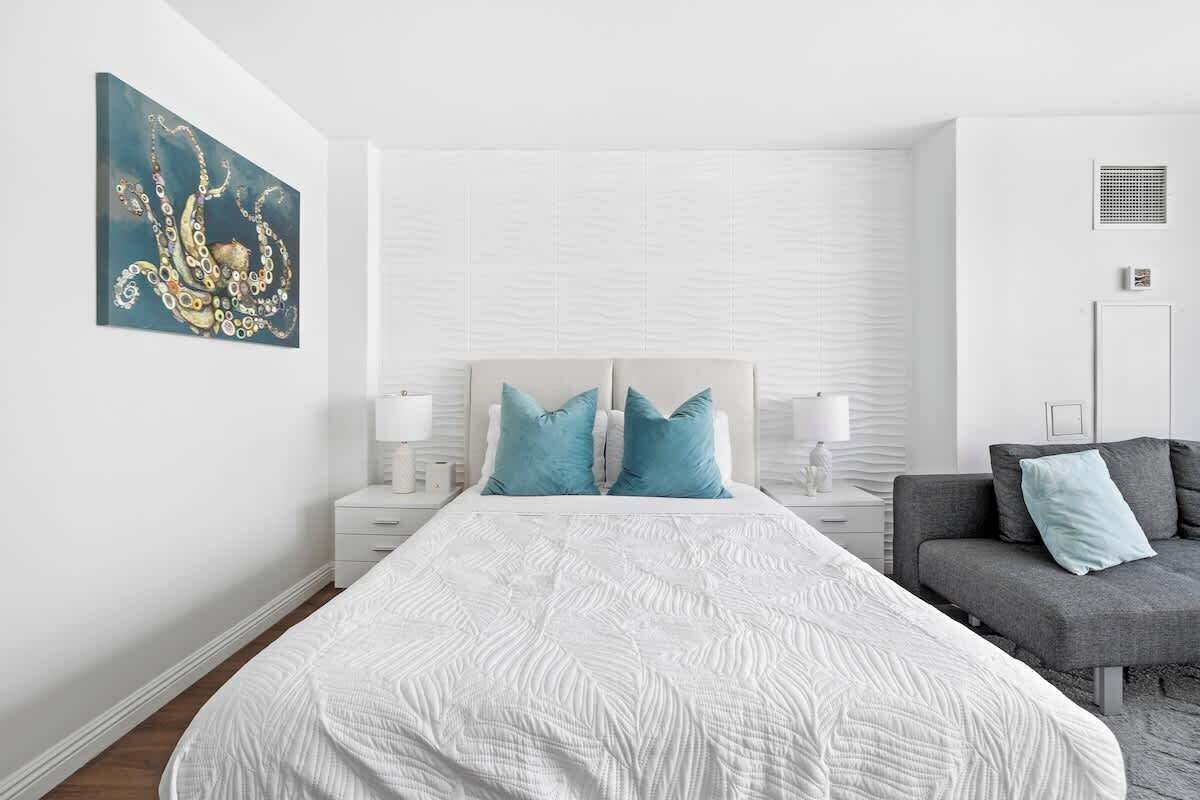 A modern bedroom with a large bed, two bedside tables with lamps, a gray sofa, and a wall-mounted artwork.