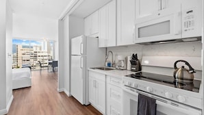 Une cuisine moderne avec des meubles blancs, une bouilloire en acier inoxydable et une vue sur le paysage urbain, visible à travers une porte ouverte.
