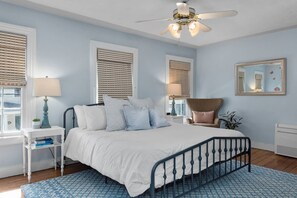 Une chambre à coucher comprenant un lit, deux tables de chevet, une chaise, un ventilateur de plafond et une fenêtre avec des stores.