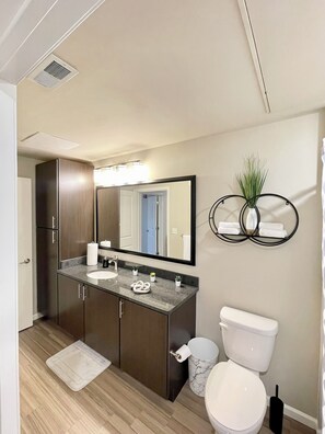 A modern bathroom with a dark wood vanity, a white sink, and a large mirror. There is a toilet with a marble trash can and a black toilet brush.