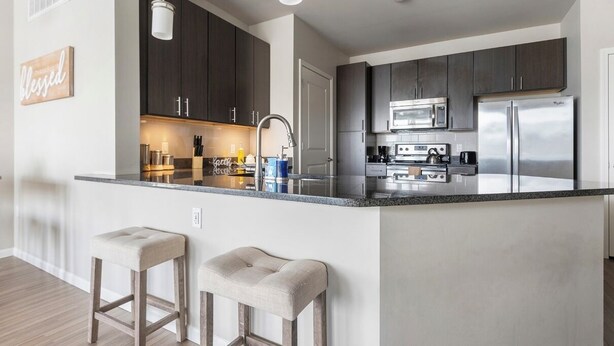 A modern kitchen with dark cabinets, stainless steel appliances, and a central island with stools. - Monthly Stays Luxe Des Peres Suite with Complimentary Starbucks Onsite!  (Des Peres)